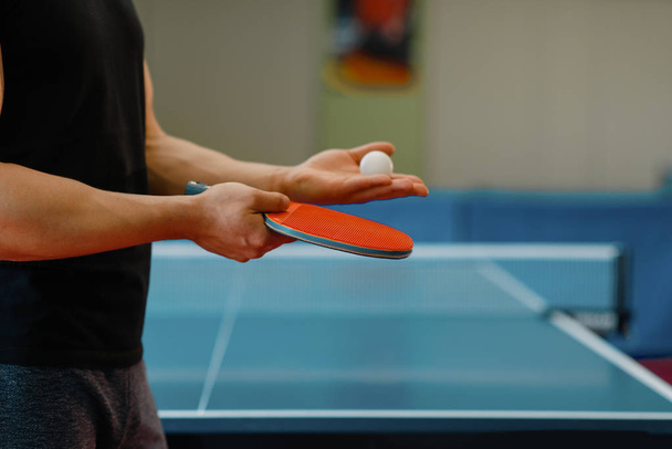 Mãos masculinas com raquete de ping pong e bola, treino dentro de casa. Homem de sportswear em pé à mesa com rede, treinando em clube de tênis de mesa
 - Foto, Imagem
