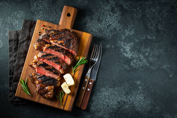 Gesneden steak Ribeye, gegrild met peper, knoflook, zout en tijm geserveerd op een houten snijplank op een donkere steen achtergrond. Bovenaanzicht met kopieerruimte. Vlakke lay - Foto, afbeelding