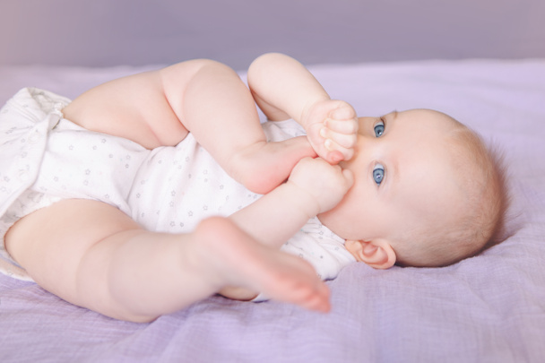Close-up portret van schattige schattige witte Kaukasische Infant kind meisje jongen met blauwe ogen liggend op bed likken zuigen vingers vuist. Baby kinderziektes. Gelukkige jeugd lifestyle en pasgeboren ontwikkeling. - Foto, afbeelding