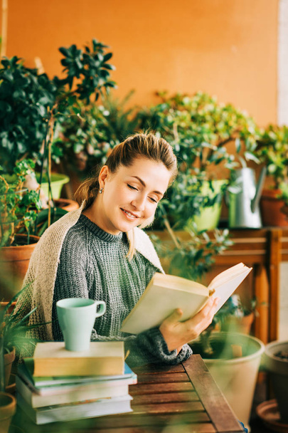 Młoda piękna kobieta relaksująca się na przytulnym balkonie, czytając książkę, ubrana w ciepłe swetry, filiżankę herbaty lub kawy na stosie książek - Zdjęcie, obraz