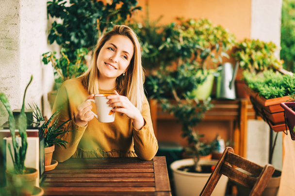 Retrato romântico de bela jovem mulher vestindo vestido de algodão marrom, relaxando na varanda entre muitas plantas verdes, segurando xícara de chá ou café
 - Foto, Imagem
