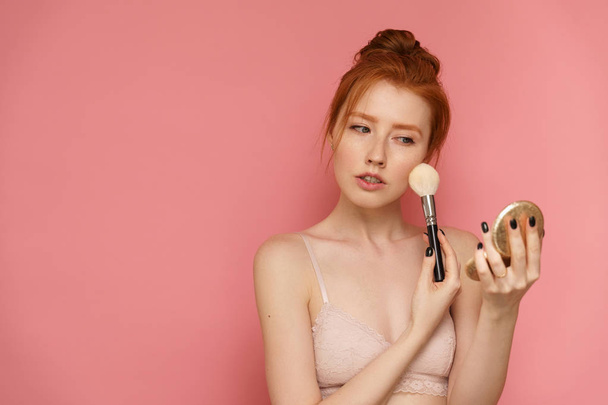 Young redhead woman with red bun makes up with a make-up brush - Foto, Imagen