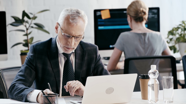 selektiver Fokus eines gutaussehenden bärtigen Mannes mit Brille, der schreibt, während er Laptop in der Nähe einer Frau im Büro benutzt   - Filmmaterial, Video