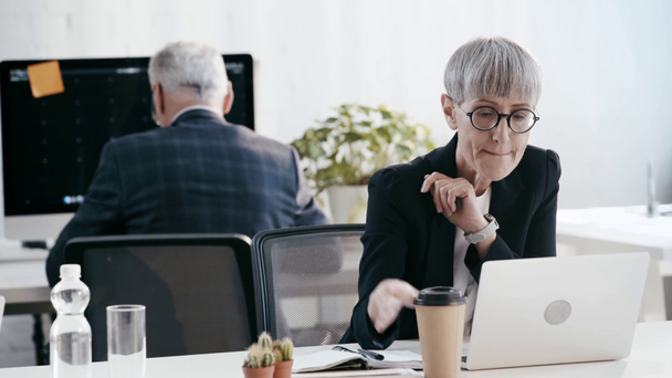 επιλεκτική εστίαση της γυναίκας στα γυαλιά ματιών χρησιμοποιώντας φορητό υπολογιστή, πίνοντας καφέ για να πάει και γράφοντας κοντά συνάδελφο στο γραφείο  - Πλάνα, βίντεο