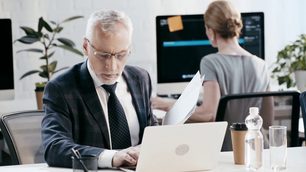 selective focus of bearded man in eye glasses holding folder with papers while using laptop and drinking coffee near coworker in office  - Footage, Video