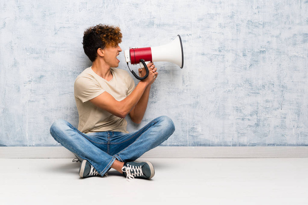 Young african american man sitting on the floor shouting through a megaphone - Photo, Image