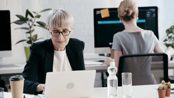 Enfoque tirón de las mujeres de negocios hablando y haciendo gestos, cuando la mujer en gafas para los ojos utilizando el ordenador portátil en la oficina
  - Metraje, vídeo
