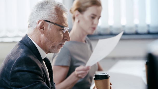 selective focus of bearded man in glasses taking paper cup and drinking coffee to go near businesswoman in office  - Video, Çekim