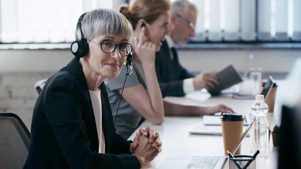 χαρούμενη γυναίκα με γυαλιά στο ακουστικό με σφικτά χέρια κοντά σε συναδέλφους στο γραφείο  - Πλάνα, βίντεο