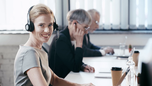 fröhliche Frau mit Headset lächelt, während sie in die Kamera in der Nähe von Kollegen im Büro schaut  - Filmmaterial, Video