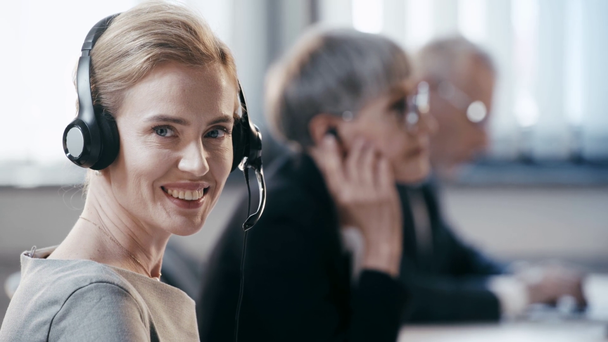 εύθυμος τηλελειτουργός στα ακουστικά κοιτάζοντας την κάμερα και χαμογελώντας κοντά σε συναδέλφους στο γραφείο  - Πλάνα, βίντεο