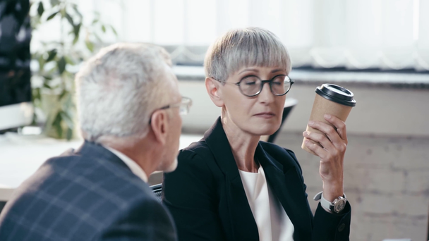 selective focus of businesswoman in glasses holding paper cup and looking at businessman talking in office  - Footage, Video