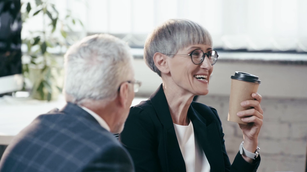 selective focus of woman in glasses holding paper cup, drinking coffee to go, looking at man and smiling in office  - Materiaali, video