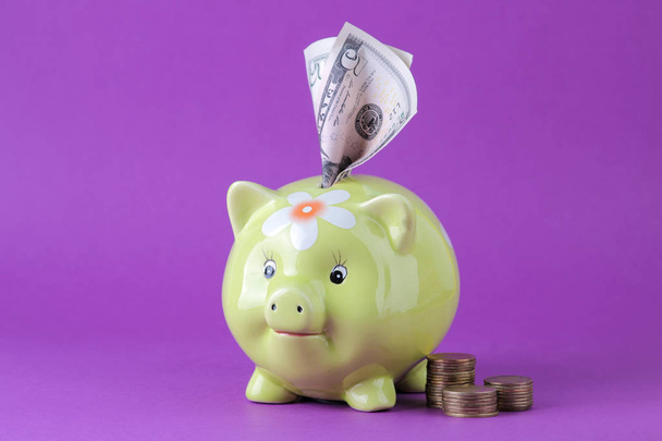 Caja de dinero de cerdo verde y dinero en un fondo lila brillante. Finanzas, ahorros, dinero. espacio para texto
 - Foto, imagen