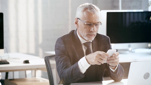 όμορφος άντρας με γένια σε γυαλιά χαμογελώντας και χρησιμοποιώντας smartphone στο γραφείο  - Πλάνα, βίντεο