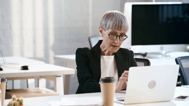 attractive woman in glasses smiling near laptop and paper cup in office  - Footage, Video