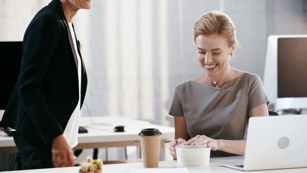 bijgesneden beeld van vrouw putting plastic container en papier Cup op tafel in de buurt van aantrekkelijke en gelukkige zakenvrouw  - Video