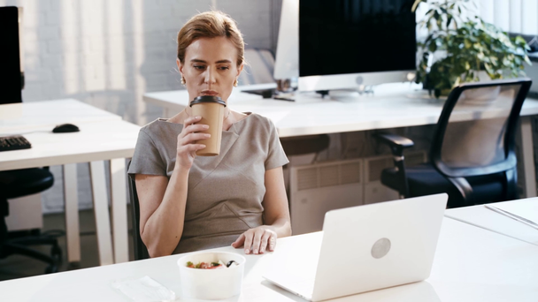 attractive businesswoman using laptop and drinking coffee to go near plastic container  - Кадры, видео