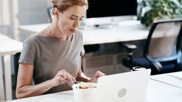mulher atraente comendo salada saborosa de recipiente de plástico perto de laptop no escritório
  - Filmagem, Vídeo