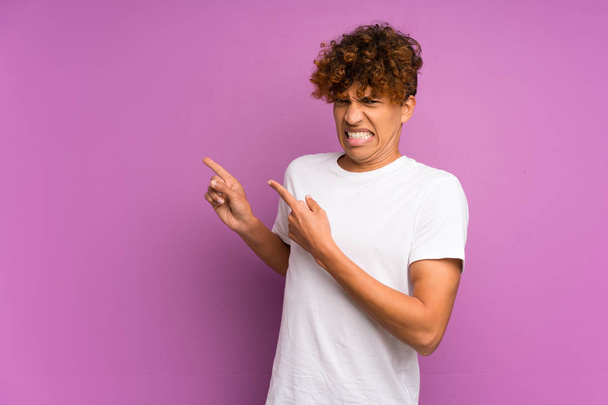 Молодий афроамериканець людина над ізольованою фіолетовий стіна злякалася і вказуючи на бік - Фото, зображення
