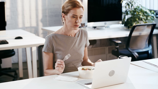 привлекательная деловая женщина берет пластиковую вилку и нож и ест вкусный салат в офисе
  - Кадры, видео