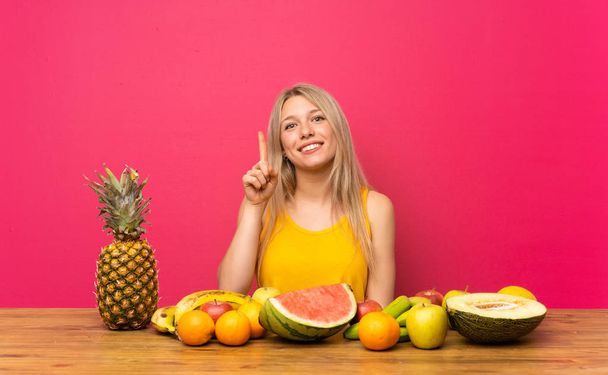 junge blonde Frau mit vielen Früchten, die mit dem Zeigefinger auf eine großartige Idee zeigt - Foto, Bild