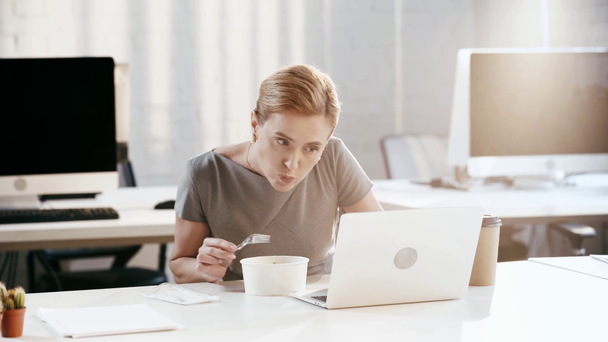 aantrekkelijke zakenvrouw met behulp van laptop, het nemen van plastic vork en het eten van salade in Office  - Video