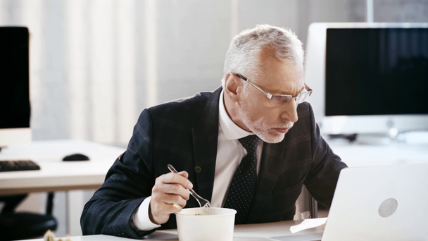 guapo hombre de negocios en gafas mirando portátil mientras come en la oficina
  - Imágenes, Vídeo