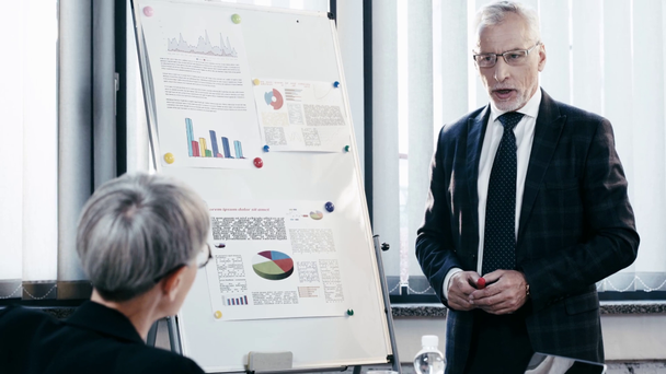 selective focus of handsome businessman in glasses talking near coworker while standing near flipchart with charts and graphs  - Metraje, vídeo
