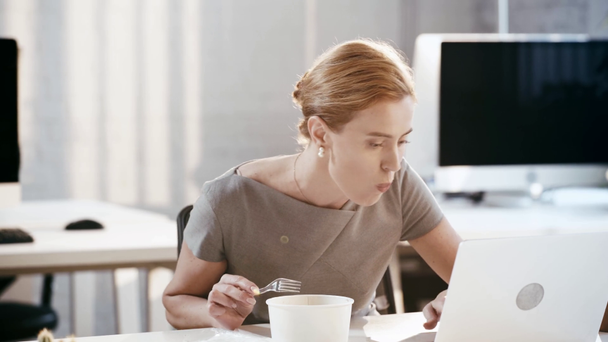 hermosa mujer de negocios usando el ordenador portátil, tomando tenedor de plástico y comer ensalada en la oficina
  - Imágenes, Vídeo