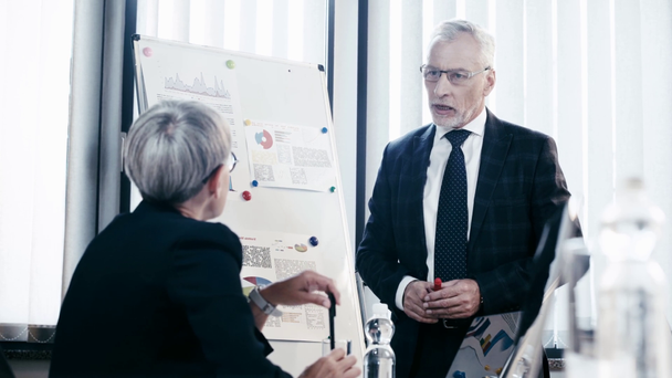 selective focus of handsome man in glasses talking near coworker while standing and gesturing near flipchart with charts and graphs  - Footage, Video