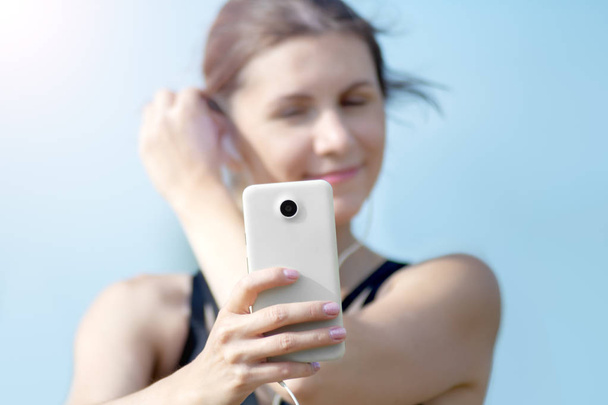 Chica sexy deportiva de fitness haciendo foto selfie en el teléfono inteligente durante el descanso de entrenamiento. Parque de la ciudad y cielo azul en el fondo
 - Foto, imagen