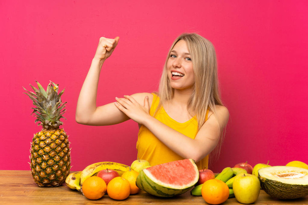 junge blonde Frau mit vielen Früchten macht starke Geste - Foto, Bild