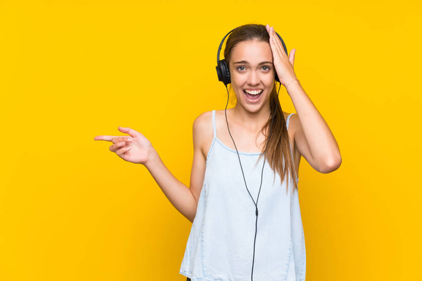 孤立した黄色い壁の上で音楽を聴いている若い女性は驚き、側面に指を指さす - 写真・画像