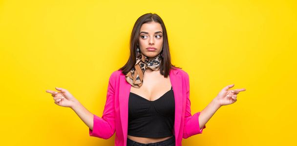 Young woman over isolated yellow background pointing to the laterals having doubts - 写真・画像
