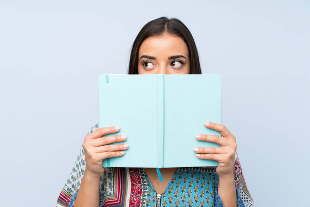 Молода жінка над ізольованою синьою стіною тримає і читає книгу
 - Фото, зображення