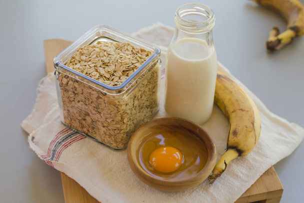 Kom met ei en banaan met granen in glazen pot met melk fles op tafel, ingrediënten voor het Cocking pannenkoeken concept  - Foto, afbeelding