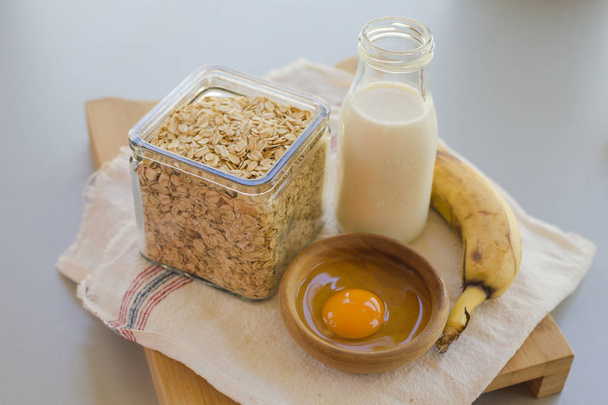 Kom met ei en banaan met granen in glazen pot met melk fles op tafel, ingrediënten voor het Cocking pannenkoeken concept  - Foto, afbeelding