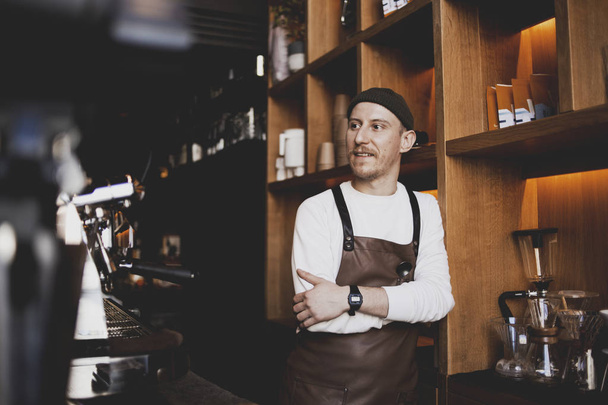 Πορτρέτο του μουσάτο ευτυχισμένο μπάρμαν στέκεται στο μοντέρνο καφέ, καφέ-μικρή επιχειρηματική ιδέα. Ένας χίπστερ μπάρμαν με μούσι. Δική του επιχείρηση, σύγχρονος επιχειρηματίας - Φωτογραφία, εικόνα