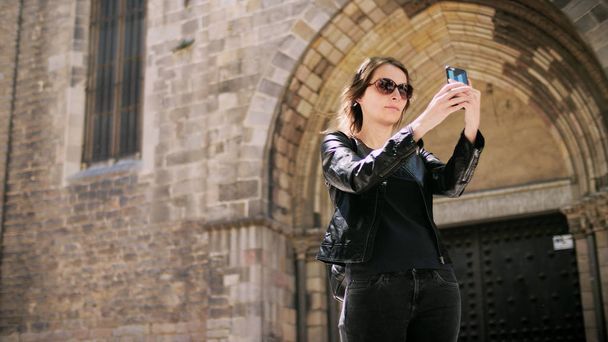 Attractive tourist woman making a selfie uses a smartfone while walking at the street. - Фото, изображение