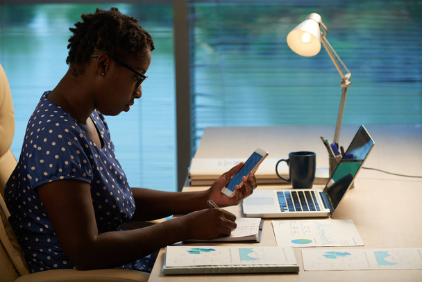 Empresária afro-americana a verificar mensagens no telemóvel e a tomar notas
 - Foto, Imagem