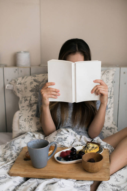 Lazy morning in warm bed. The girl has breakfast in the morning in her room. Beautiful and cute girl reading book in the winter a cozy atmosphere. Eating fresh berries, sweet piece of cake, hot tea. - Foto, imagen