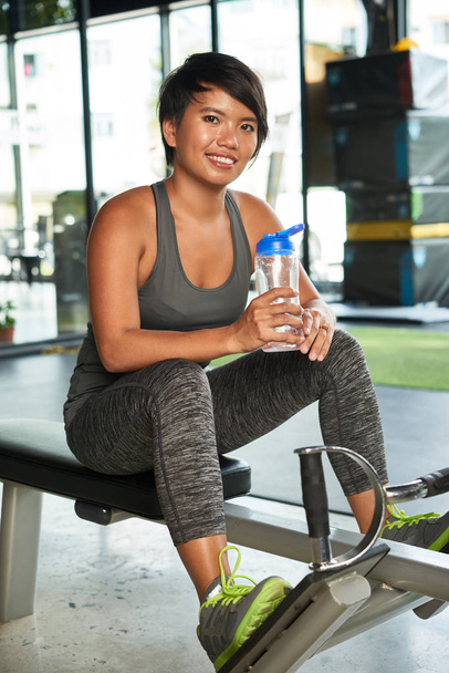 Портрет молодої жінки, що сидить на спортивному обладнанні з пляшкою води і посміхається в камері в клубі здоров'я
 - Фото, зображення