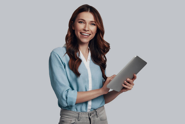 Vizsgálva az új tablettát. Vonzó fiatal nő dolgozik a digitális tabletta, és mosolyogva állva ellen szürke háttér - Fotó, kép