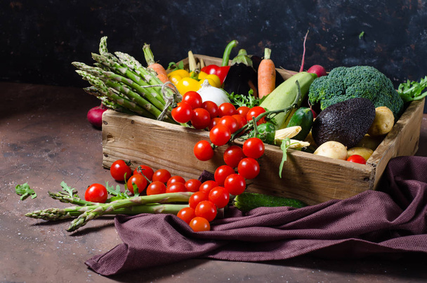Caixa com legumes de fazenda em um fundo escuro. Lugar para SMS. Tomates cereja, espargos, brócolis, pimentas, berinjelas, abobrinhas, cenouras e cebolas
. - Foto, Imagem