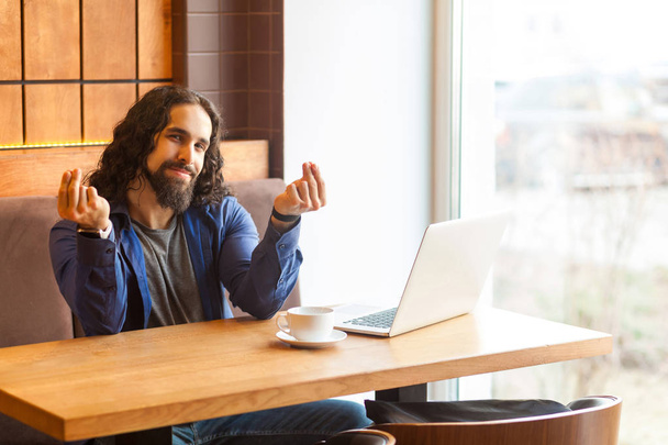 handsome young adult man freelancer in casual style sitting in cafe with laptop and showing cash gesture while looking at camera - Foto, Imagen