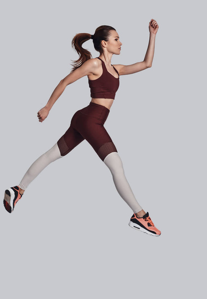 Latanie wysoko. Pełna długość atrakcyjna młoda kobieta w odzieży sportowej skoki podczas ćwiczeń na szarym tle - Zdjęcie, obraz