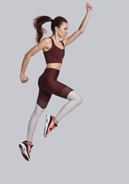 Συνέχισε να κινείσαι! Πλήρες μήκος της ελκυστική νεαρή γυναίκα σε αθλήματα ρούχα άλμα κατά την άσκηση σε γκρίζο φόντο - Φωτογραφία, εικόνα