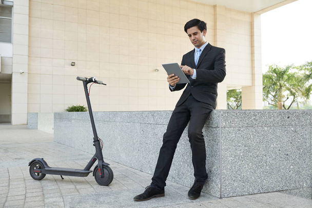 Молодой бизнесмен в костюме работает в Интернете на цифровом планшете, сидя в городе с электрическим скутером
 - Фото, изображение