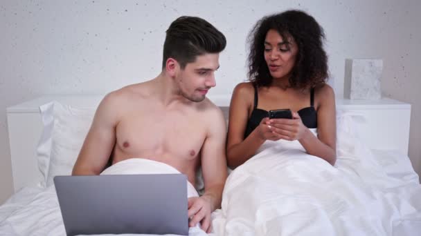 Smiling loving multiethnic couple using laptop computer and smartphone while lying in bed - Filmati, video